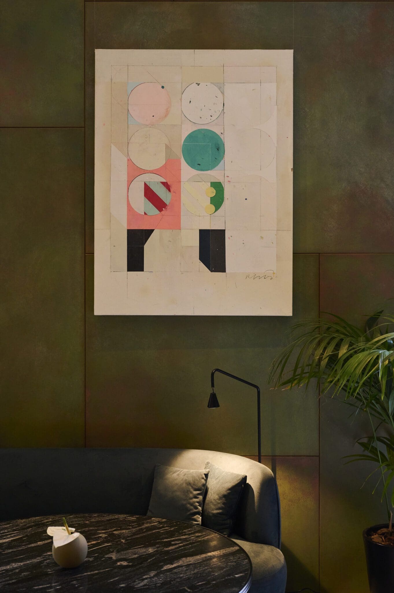 Abstract geometric artwork with pastel circles and rectangles hangs on a green wall above a black sofa, with a small table, vase, lamp, and potted plant nearby.
